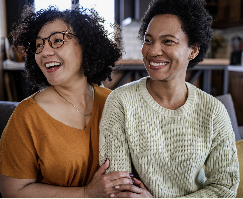 Two women happily smiling and holding each other in a cozy indoor setting.